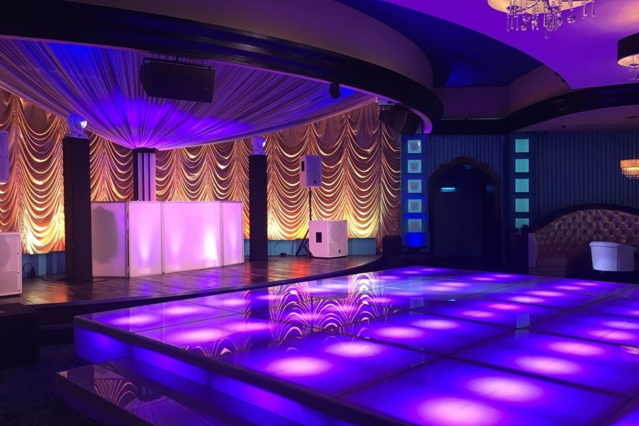 A vibrant event setup featuring a glowing purple LED dance floor, gold draped backdrop, and ambient lighting.