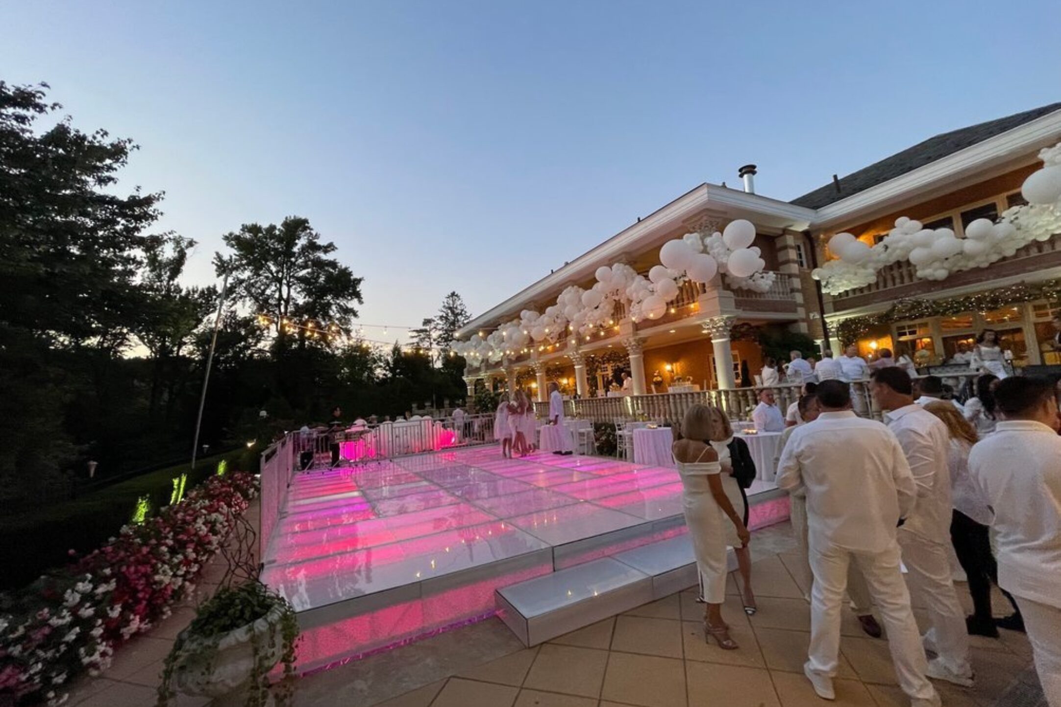 An elegant outdoor celebration with a glowing glass dance floor, surrounded by white floral decorations and balloons, creating a dreamy ambiance.