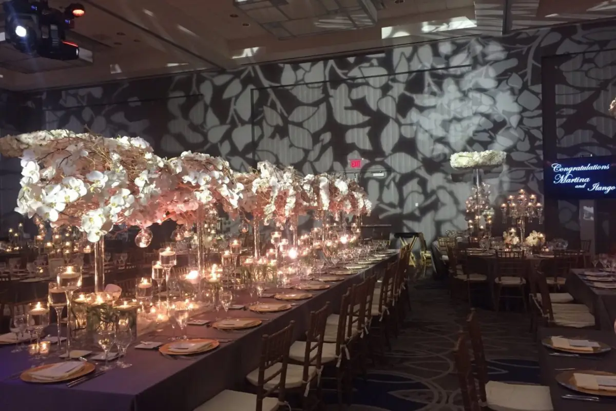 Sophisticated indoor event setup with intricate floral centerpieces, candlelit tables, and patterned wall lighting.